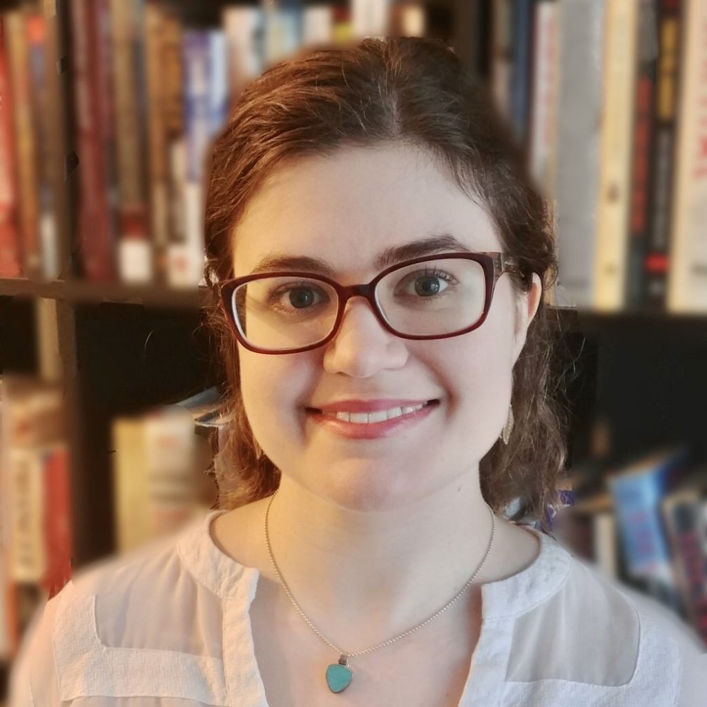 Headshot of Sarah Brand. Sarah is standing in front of a bookshelf. She has wavy brown hair and glasses. She is wearing a white blouse and a turquoise necklace.